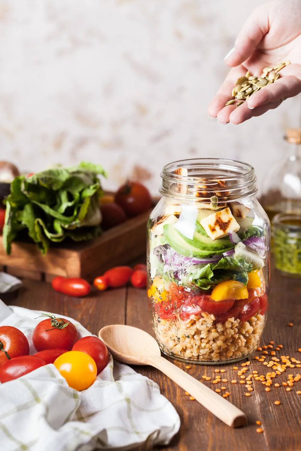 A hand sprinkling seeds over a jar layered with grains, vegetables and fresh produce